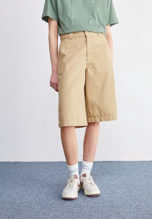 Person iført lysebrune knælange shorts, grøn skjorte, hvide sokker med blå striber og hvide sneakers står på gråt tæppe.