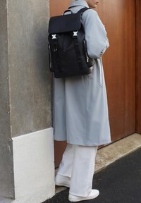 Schwarzer Rucksack mit Klappenverschluss, silbernem Zubehör, seitlichen Reißverschlüssen und verstellbaren Trägern, kombiniert mit einem hellgrauen Mantel und weißen Hosen.