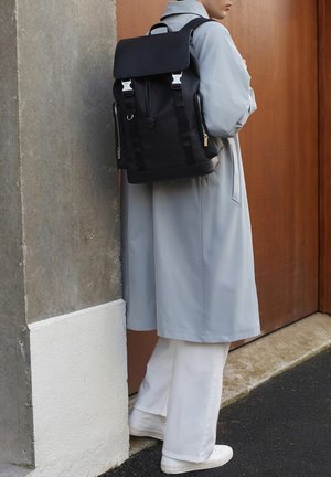 Schwarzer Rucksack mit Klappenverschluss, silbernem Zubehör, seitlichen Reißverschlüssen und verstellbaren Trägern, kombiniert mit einem hellgrauen Mantel und weißen Hosen.