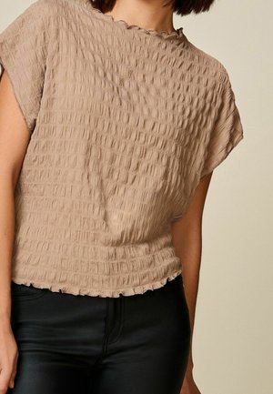 Femme portant un haut beige texturé à manches courtes avec un ourlet ondulé et un pantalon noir, vue des épaules aux hanches sur un fond uni.