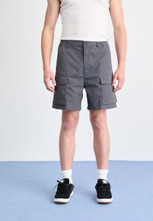 Graue Cargo-Shorts mit mehreren Vordertaschen, einem elastischen Bund und geradem Beinanschnitt. Tragen mit schwarzen Turnschuhen und weißen Socken.