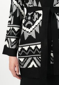 Pletený cardigan s geometrickým vzorem v černé, bílé a šedé barvě, s opaskem v pase a žebrovanými manžetami pro zvýšení textury.