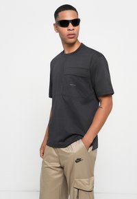 Mand iført sorte solbriller, mørkegrå kortærmet skjorte med brystlomme og beige Nike cargo bukser med hænderne i lommerne.