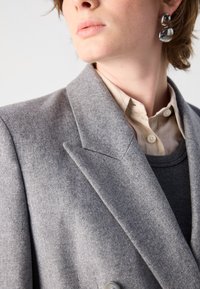 Grijze wollen blazer met geprofileerde revers, gestructureerde stof en dubbele knoopsluiting. Draag het over een grijze trui en een beige shirt met knoopdetails.