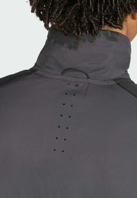 Person with dark curly hair wearing a high-collar dark jacket with vertical ventilation holes and a small fabric loop at the back.