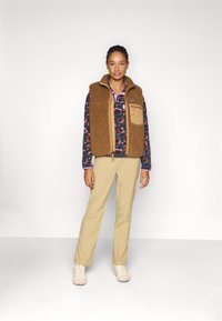 Gilet en polaire marron sur une chemise à motifs, pantalon beige et chaussures claires. Il possède des poches et un col haut. Design simple.