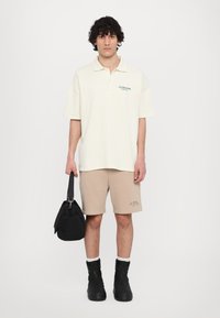 Polo shirt crème avec texte vert, shorts beiges avec un imprimé signature, et bottes noires à zip. Sac à main noir à la main.