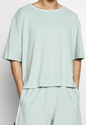 Mężczyzna noszący jasnoszary, krótki rękaw z frotte koszulę oraz dopasowane szorty, z rękami w kieszeniach, przedstawiony od szyi do połowy uda.