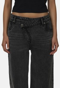 Czarne dżinsy typu wide-leg z unikalnym skośnym pasem i zapięciem na guzik, z delikatnym wyblaknięciem i kieszeniami po obu stronach.