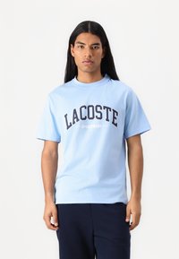 Světle modré bavlněné tričko s výrazným černým nápisem "LACOSTE SPORTSWEAR" přes hrudník, kulatým výstřihem a krátkými rukávy.