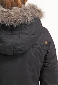 Sort parka-jakke med en faux fur-hættebesætning, der har et holdbart stof, synlige syninger og knapdetaljer på skuldrene.