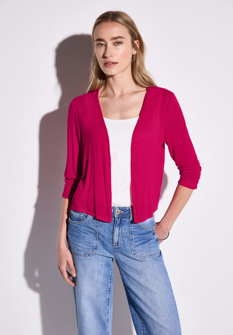 Cardigan fuchsia en tissu léger avec une coupe décontractée, devant ouvert et manches trois-quarts, associé à un jean bleu clair.