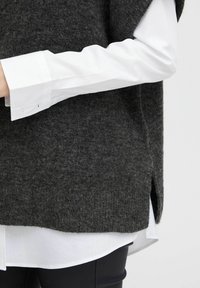 Grauer Wollpullover mit lockerer Passform, versehen mit einem seitlichen Schlitz, über einem weißen Hemd mit Knopfmanschetten und glatter Textur getragen.