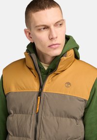 Padded vest in tan and brown, featuring a zippered front, stand-up collar, and a logo. Worn over a green hoodie. Textured fabric visible.