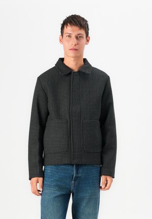 Chaqueta de patrón de espiga en negro y gris con cuello, dos bolsillos frontales y corte relajado, llevada sobre jeans azules.