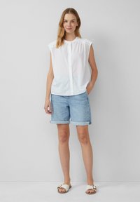 Chemise sans manches blanche à boutons, shorts en denim bleu clair retroussés, et sandales blanches à enfiler avec une boucle dorée. Fond uni.