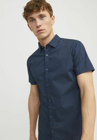 Camicia a maniche corte di un blu scuro con un motivo a pois. Presenta un colletto a punta, tessuto morbido e una forma aderente.