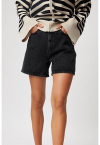 Persona vestida con pantalones cortos de mezclilla negros y un suéter de manga larga con cremallera de rayas beige y negras, con las manos en los bolsillos.