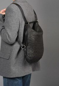 Zwarte leren rugsack met geweven textuur, verstelbare schouderriem en zilverkleurige hardware, gedragen over een grijze blazer en blauwe jeans.