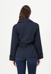 Marineblauwe trenchcoat met een ceintuurband, brede kraag en knoopmanchetten, gemaakt van een gladde, gestructureerde stof. Achteraanzicht.