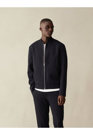 Veste de bombardier marine avec une fermeture éclair à l'avant, ourlet et poignets côtelés, deux poches latérales, portée par-dessus une chemise blanche. Tissu lisse, coupe ajustée.