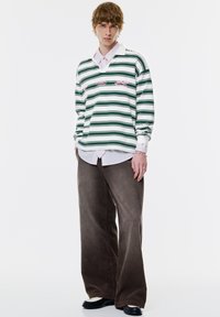 Zielono-biała w paski koszula z długim rękawem z kołnierzykiem, nałożona na jasnoróżową koszulę z guzikiem, brązowe szerokie spodnie z kordurowego materiału.