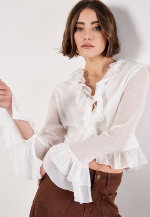 Femme aux cheveux bruns courts portant un chemisier blanc transparent avec des volants et un pantalon marron taille haute, posant sur un fond clair.