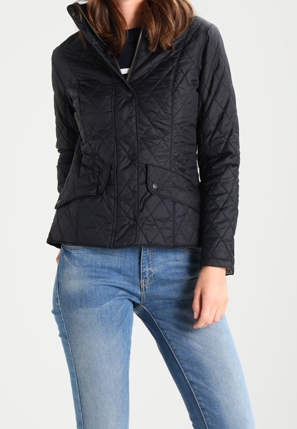 Femme portant une veste matelassée noire avec des poches avant et un jean bleu, debout devant un fond clair uni.