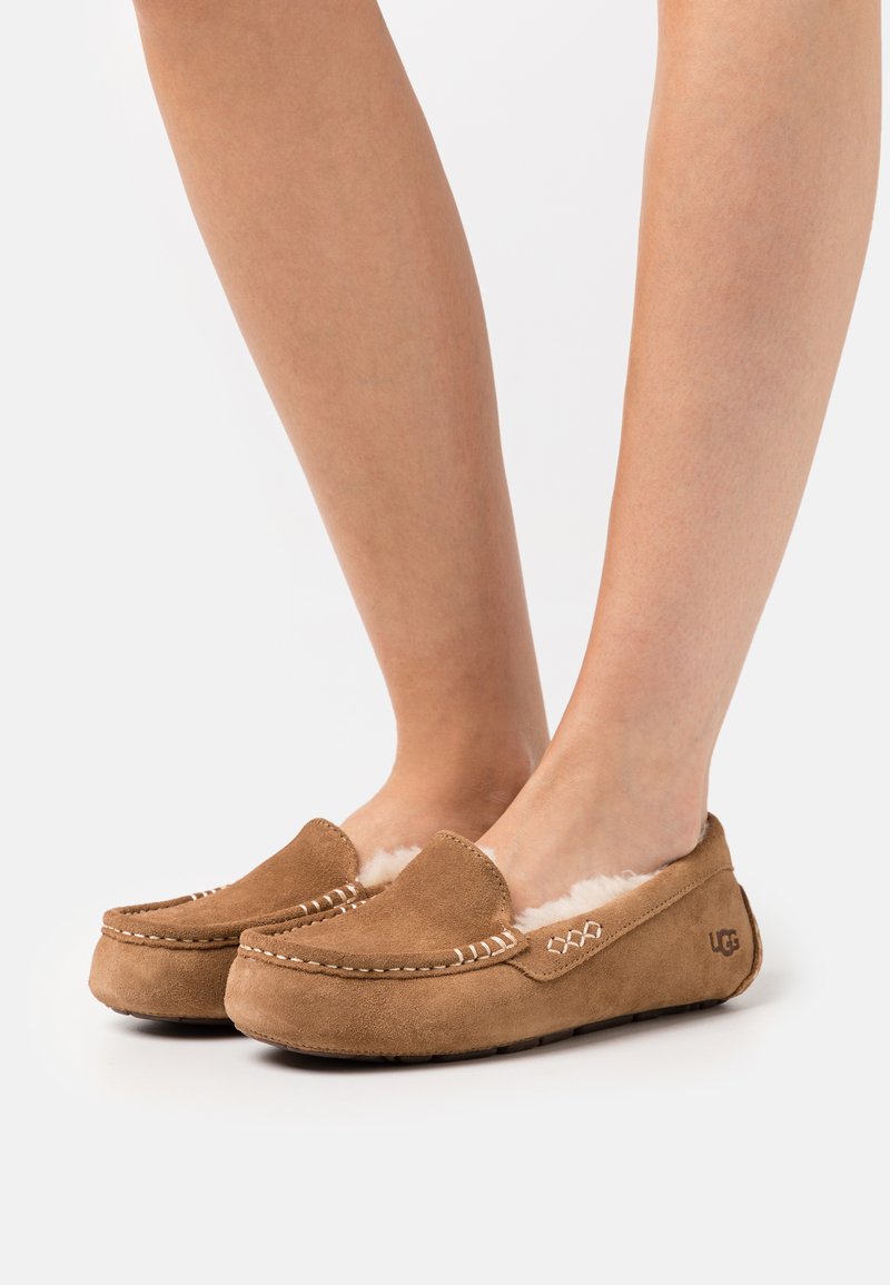 Bruine suède moccasin slippers met witte stikdetails, zachte binnenvoering en UGG-branding aan de zijkant. Plat uiterlijk met een ronde neus.