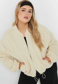 Kremowa, puszysta kurtka bomberka z prążkowanym kołnierzem, zamkiem z przodu i ściąganym dołem. Oferuje luźny krój i fakturowany materiał.