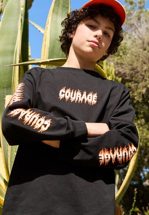 Camiseta negra de manga larga con un gráfico de texto de llamas naranjas que dice "CORaje" en el frente y las mangas. El modelo está de pie contra una planta verde.