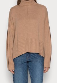 Beżowy sweter z golfem, o prążkowanej fakturze, w oversize'owym kroju oraz z bocznymi rozcięciami. Połączony z niebieskimi dżinsami. Posiada długie rękawy i luźny design.