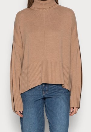 Sandfarbener Rollkragenpullover mit geripptem Muster, oversized Schnitt und seitlichen Schlitzen. Getragen zu blauen Jeans. Verfügt über lange Ärmel und ein lockeres Design.