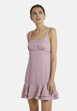 Frau trägt ärmelloses pinkfarbenes Kleid mit quadratischem Ausschnitt und geschichtetem Rüschensaum, schaut leicht nach oben und zur Seite.