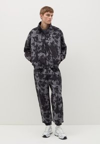 Tuta a motivi neri e grigi, dotato di giacca con zip e polsini elasticizzati e pantaloni abbinati con vita elasticizzata. Sneakers bianche.