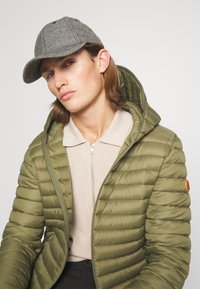 Olijfgroene, puffjacket met een horizontaal doorgestikt ontwerp, gecombineerd met een lichtbeige, met rits afgesloten top en een grijze, gestructureerde pet.
