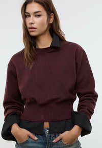 Giovane donna che indossa un maglione corto bordeaux sopra una camicia nera a righe con i polsini arrotolati, abbinata a jeans blu, con le mani nelle tasche.