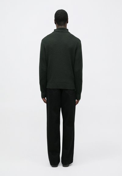 Mörkgrön stickad tröja med hög krage, kombinerad med svarta byxor med vida ben. Tyget verkar mjukt med en slät yta.