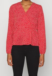 Blusa rossa con scollo a V, maniche lunghe e taglio peplum. Presenta un piccolo motivo a cuori bianche e polsini delle maniche arricciati.