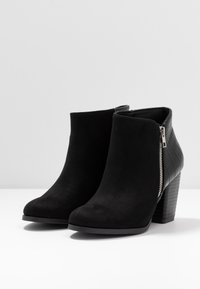 Et par sorte ruskind ankelstøvler med lynlås i siden og tekstureret læderhæl og bagpanel, sat mod en ensfarvet hvid baggrund.
