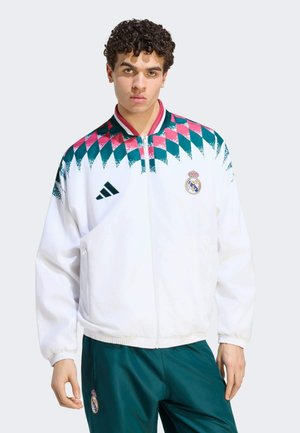 Jonge man draagt een wit Real Madrid-jack met een turquoise en roze diamantenpatroon en bijpassende groene sportbroek tegen een lichte achtergrond.