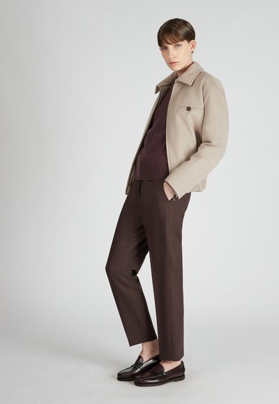 Giacca in lana beige, maglione a maglia viola scuro, pantaloni marroni e mocassini scuri. La giacca ha un colletto e dettagli con bottoni; trame lisce in tutto.