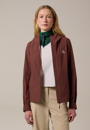 Giovane donna che indossa una giacca con cerniera color borgogna sopra una maglietta bianca e pantaloni beige, con una sciarpa verde intorno al collo, su uno sfondo semplice.