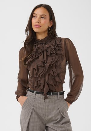 Femme portant un chemisier marron transparent à volants, un pantalon gris à carreaux taille haute, une ceinture noire et des boucles d'oreilles créoles, les mains dans les poches.