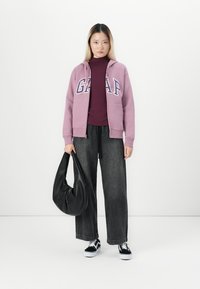 Vrouw in een mauve GAP-hoodie, bordeaux top, wijde zwarte jeans, zwart-witte sneakers, met een grote zwarte handtas voor een witte achtergrond.