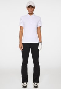 Witte poloshirt met korte mouwen en subtiele textuur, zwarte golfbroeken, witte handschoen en zwart-witte golfschoenen, met een beige pet.
