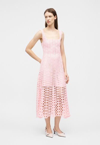 Femme portant une robe midi rose sans manches avec un motif de découpes circulaires et des escarpins blancs à bride arrière, debout devant un fond clair uni.