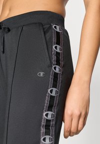 Μαύρα τζόγκερ με ριμπ ύφασμα στη μέση και κορδόνι, με πλαϊνές ρίγες με γκρι σχέδια. Ομαλή υφή και έμβλημα στην περιοχή του μηρού.