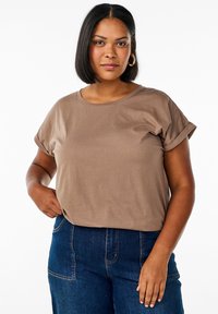 Braunes T-Shirt mit kurzen Ärmeln und umgeschlagenen Ärmeln, aus weichem Baumwollmaterial gefertigt. Kombiniert mit dunkelblauen Jeans mit Taschen und Nähten.
