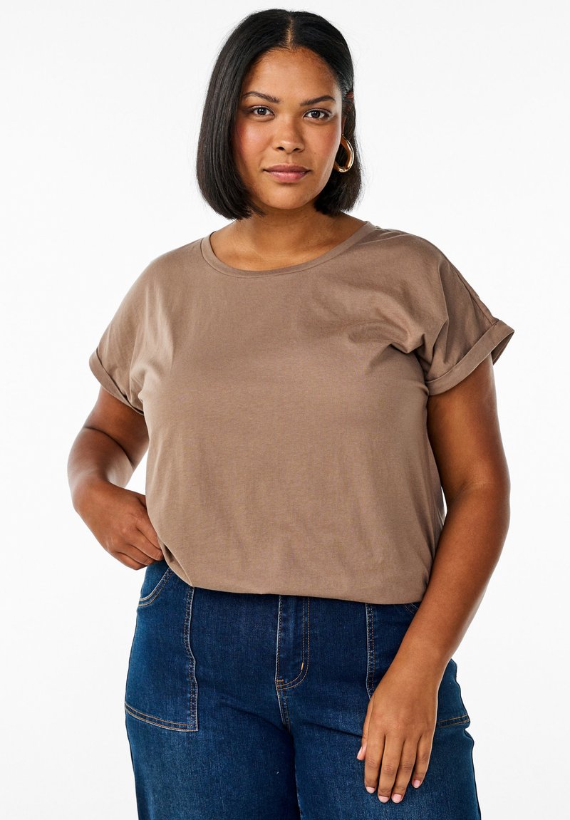 Braunes T-Shirt mit kurzen Ärmeln und umgeschlagenen Ärmeln, aus weichem Baumwollmaterial gefertigt. Kombiniert mit dunkelblauen Jeans mit Taschen und Nähten.